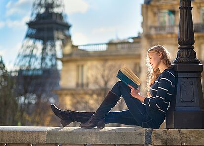 Studentin mit Buch sitzt auf einem Brückengeländer mit Blick auf den Eiffelturm
